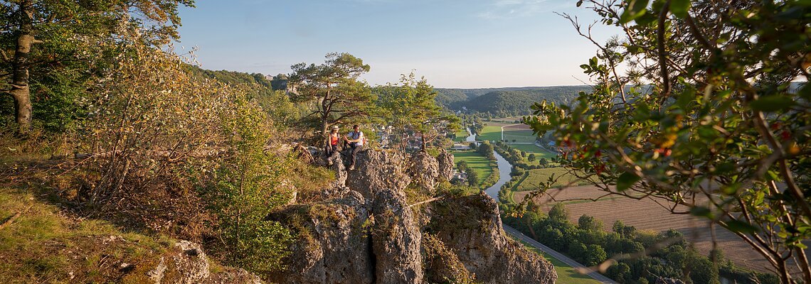 Ein Wanderpärchen, etwas entfernt, steht links auf einem Felsen an der Arnsberger Leite, die gut zu sehen ist. Dahinter erstreckt sich der Blick ins Tal. Der Himmel erstrahlt wolkenlos.