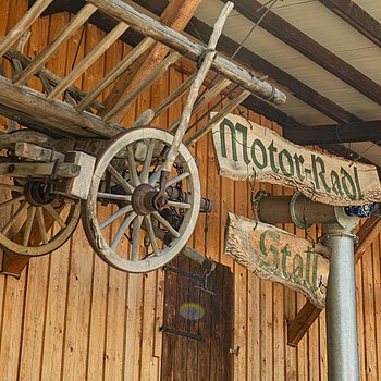 Holzwagenrad und Holzschilder mit der Aufschrift "Motor-Radl Stall" an einer Holzwand.