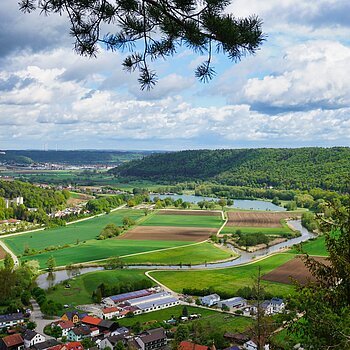 Landschaft mit Feldern, Fluss und Waldhügeln unter bewölktem Himmel, im Vordergrund ein Dorf.