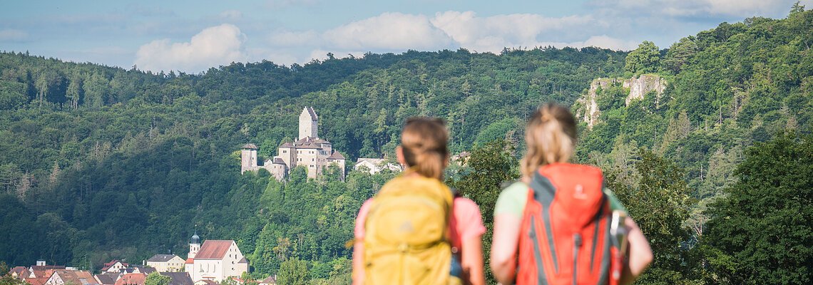 Zwei Personen mit Rucksäcken blicken auf eine Burg in einer bewaldeten Landschaft.
