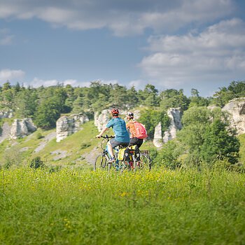 Zwei Personen fahren mit Fahrrädern auf einem grasbewachsenen Feld vor einer Felslandschaft.