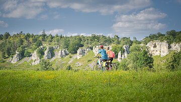 Zwei Personen fahren mit Fahrrädern auf einem grasbewachsenen Feld vor einer Felslandschaft.