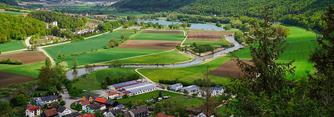 Landschaft mit Feldern, Fluss und Waldhügeln unter bewölktem Himmel, im Vordergrund ein Dorf.