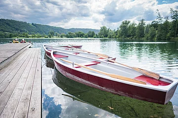 Zwei Ruderboote sind an einem Holzsteg festgemacht, umgeben von einem See und bewaldeten Hügeln.