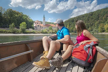 Paar sitzt in einem Boot auf einem Fluss und blickt auf ein Gebäude mit Turm und Wald im Hintergrund.