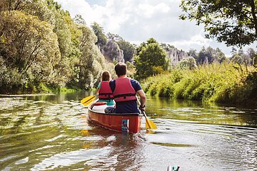 Zwei Personen mit roten Schwimmwesten paddeln in einem roten Kanu auf einem Fluss, umgeben von grüner Natur.
