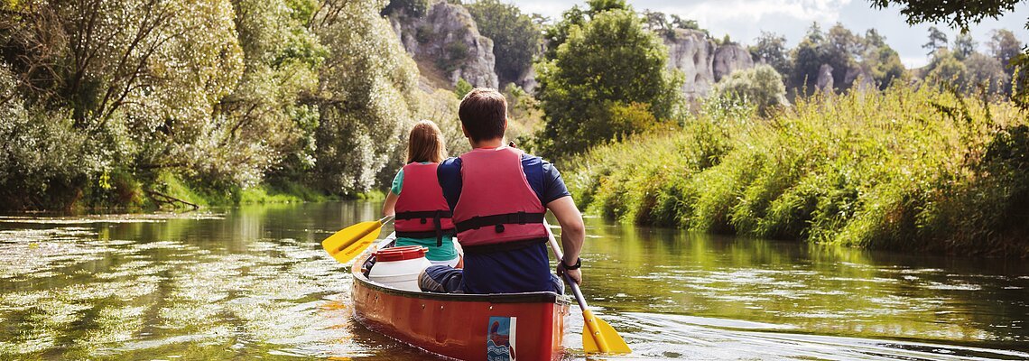 Zwei Personen mit roten Schwimmwesten paddeln in einem roten Kanu auf einem Fluss, umgeben von grüner Natur.