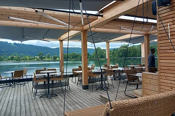 Überdachte Terrasse mit Tischen und Stühlen an einem See, im Hintergrund Bäume und bewaldete Hügel.