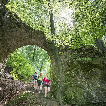 Vier Wanderer gehen einen Waldweg mit Steintreppe unter einem natürlichen Felsbogen hinab.