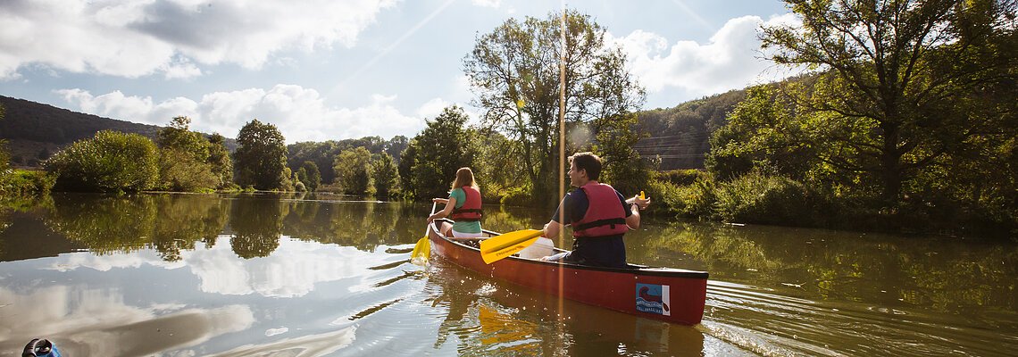 Zwei Personen paddeln in einem roten Kanu auf einem ruhigen Fluss bei sonnigem Himmel mit Wolken.