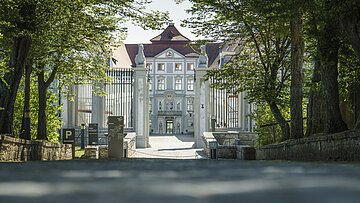 Eingangstor zu einem historischen Gebäude mit Bäumen und Schildern am Wegesrand bei Tageslicht.