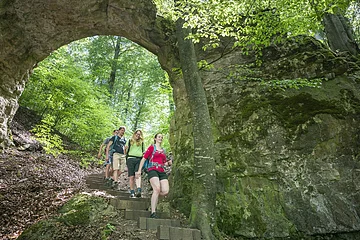 Vier Wanderer gehen einen Waldweg mit Steintreppe unter einem natürlichen Felsbogen hinab.