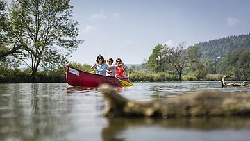Drei Personen paddeln in einem roten Kanu auf einem ruhigen Fluss mit Bäumen und Hügeln im Hintergrund.