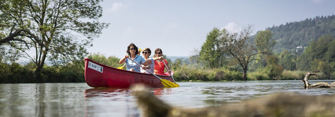 Drei Personen paddeln in einem roten Kanu auf einem ruhigen Fluss mit Bäumen und Hügeln im Hintergrund.