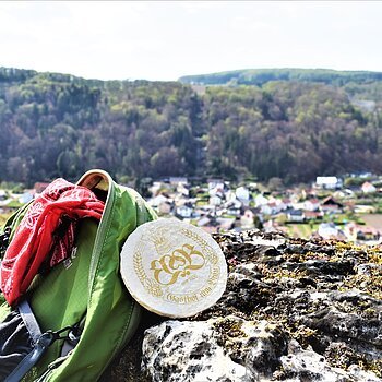 Hotel zum Bräu Wanderung rund um Enkering Grüner Rucksack mit rotem Tuch und rundem Bierdeckel auf Felsen, im Hintergrund Dorf und bewaldete Hügel.