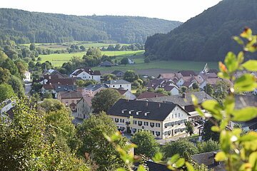 Blick auf ein Dorf mit Häusern, Bäumen und Feldern, umgeben von bewaldeten Hügeln an einem sonnigen Tag.