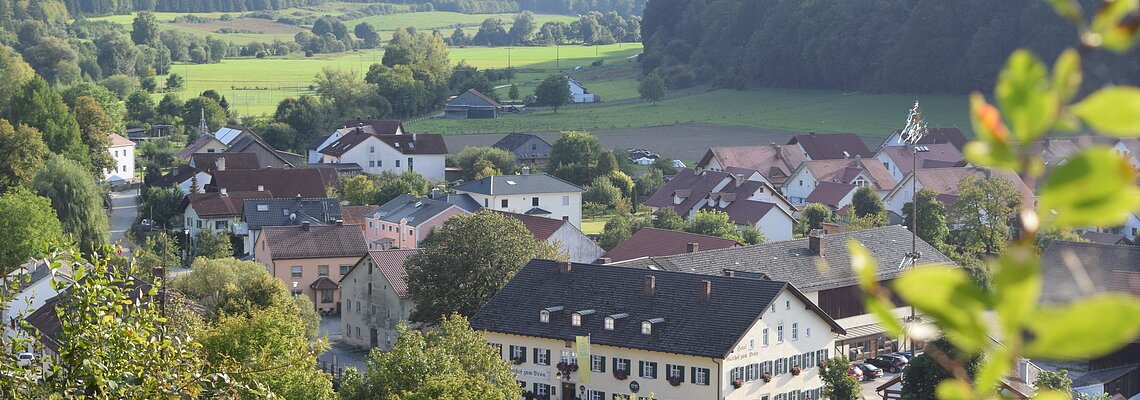 Blick auf ein Dorf mit Häusern, Bäumen und Feldern, umgeben von bewaldeten Hügeln an einem sonnigen Tag.