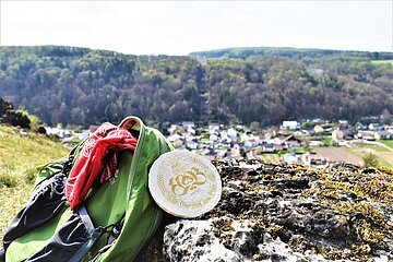 Grüner Rucksack mit rotem Tuch und rundem Bierdeckel auf Felsen, im Hintergrund Dorf und bewaldete Hügel.