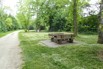 Unter dem Bäumen steht eine gemütliche Sitzgelegenheit aus Holz. Links führt ein geschotterter Weg entlang.