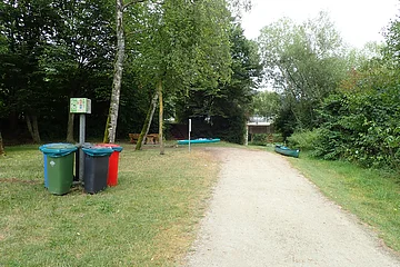 Waldweg mit Mülltonnen, Bänken, Kanus und Brücke im Hintergrund, umgeben von Bäumen und Grasfläche.