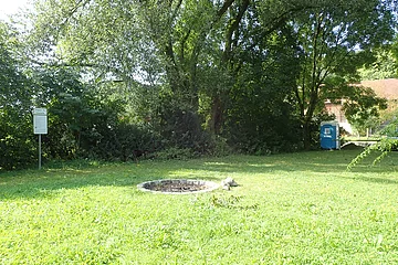 Grüne Wiese mit Feuerstelle, Bäumen, einem Hinweisschild links und einem blauen mobilen Toilettenhäuschen rechts.