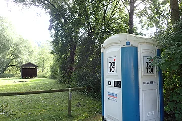 Mobiles Toilettenhäuschen mit blau-weißer Außenwand steht am Waldrand auf einer Wiese neben einem Holzzaun.