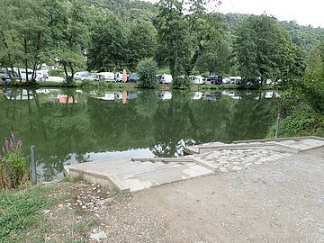 Ufer mit Treppe an einem ruhigen See, gegenüber Bäume und Wohnwagen auf einer Wiese.