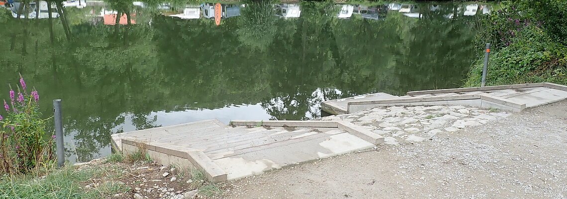 Ufer mit Treppe an einem ruhigen See, gegenüber Bäume und Wohnwagen auf einer Wiese.