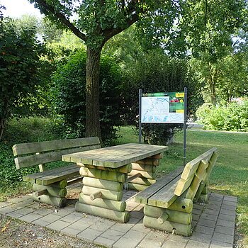 Bootsrastplatz Ilbling Holztisch mit zwei Bänken auf gepflastertem Platz unter Baum, im Hintergrund Infotafel und Grünfläche.