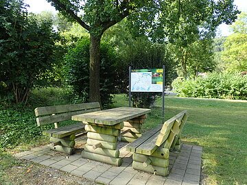 Holztisch mit zwei Bänken auf gepflastertem Platz unter Baum, im Hintergrund Infotafel und Grünfläche.