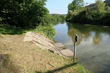 Ufer mit Stufen aus Beton an einem ruhigen Fluss, umgeben von Bäumen und Grasflächen unter blauem Himmel.