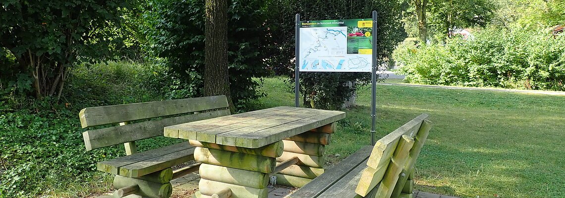 Holztisch mit zwei Bänken auf gepflastertem Platz unter Baum, im Hintergrund Infotafel und Grünfläche.