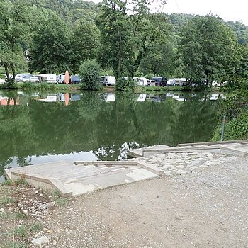 Anlegestelle Kratzmühle Ufer mit Treppe an einem ruhigen See, gegenüber Bäume und Wohnwagen auf einer Wiese.